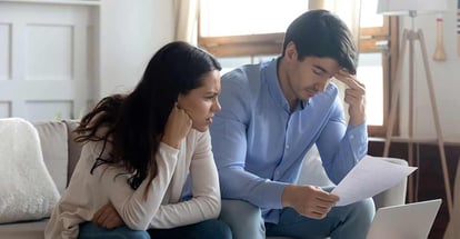 Concerned worried young married couple sitting on sofa at home studying document