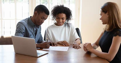 Excited African American couple sit at desk with female agent and sign agreement