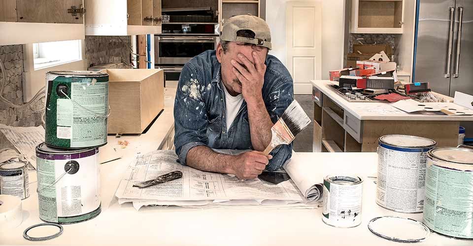 Exhausted and frustrated house painter in messy clothes with face in hands and holding wet paintbrush in fixer upper home kitchen remodel
