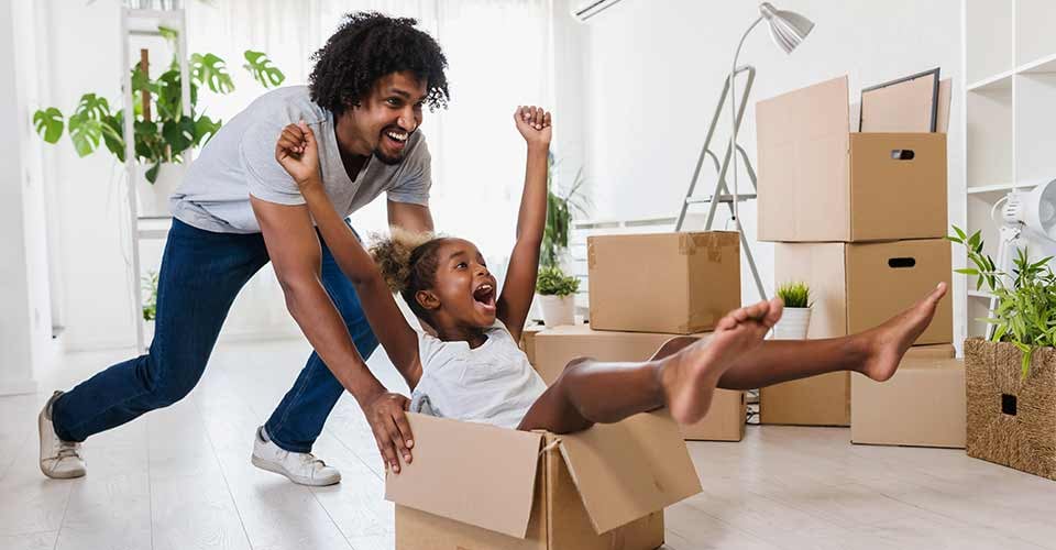 Father push cute little daughter sitting inside of carton box having fun riding in living room