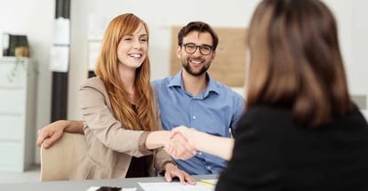 Happy young couple meeting with a broker in her office leaning over the desk to shake hands