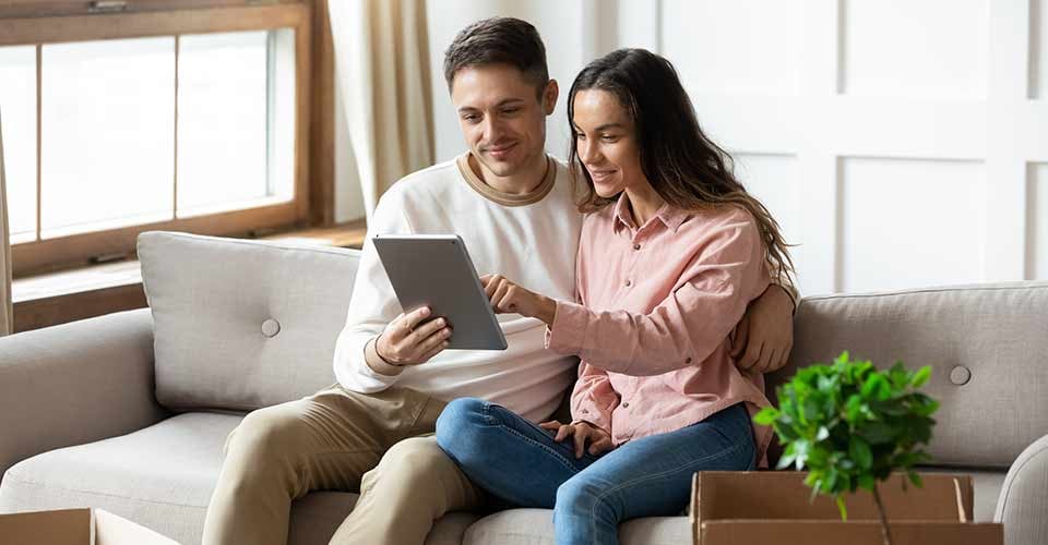 Happy young couple reading document on tablet together sitting on couch in living room