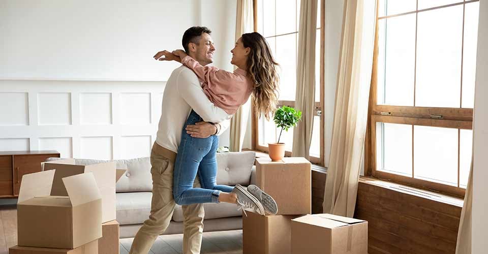 Happy young husband lifting excited wife celebrating moving day with cardboard boxes