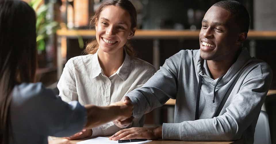 Happy young interracial couple shake hand of broker to take mortgage loan