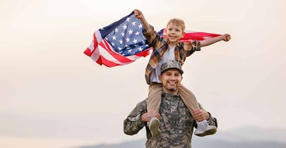 Military father carrying happy little son with american flag on shoulders