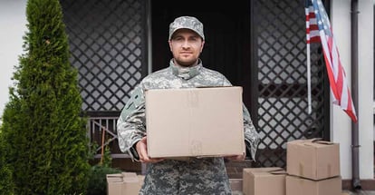 Military serviceman with cardboard box standing near house with american flag