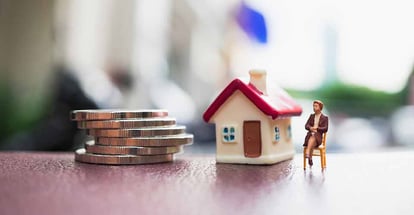 Miniature person sitting in front of house with pile of coins